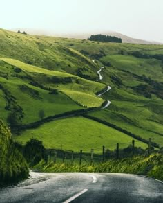 an empty road in the middle of a lush green valley with hills and fields on either side