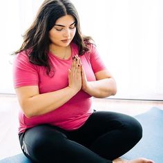 a woman in pink shirt and black pants sitting on yoga mat with her hands together