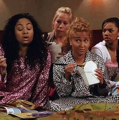 four women sitting on a bed eating food and drinking coffee while looking at the camera