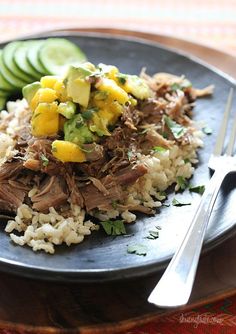 a black plate topped with rice and meat next to cucumber slices on a table