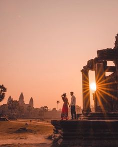 two people standing in front of an ancient temple at sunset with the sun setting behind them