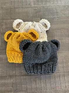 three knitted hats are laying on a wooden surface, with one bear's head in the middle