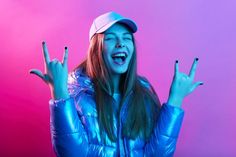 a woman wearing a blue jacket and hat making the peace sign with her hands while standing in front of a pink background