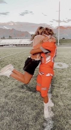 two people are hugging in the middle of a soccer field with mountains in the background