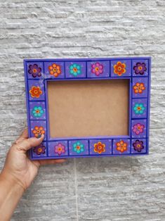 a person holding up a purple frame with colorful flowers on the front and sides, against a stone wall