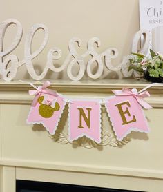 a pink and gold first birthday banner hanging from a mantel