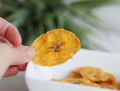 a person holding up a piece of food in front of a white bowl filled with bananas