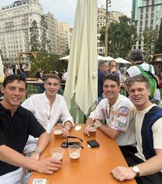 three men sitting at a table with beer glasses in front of them and people standing around