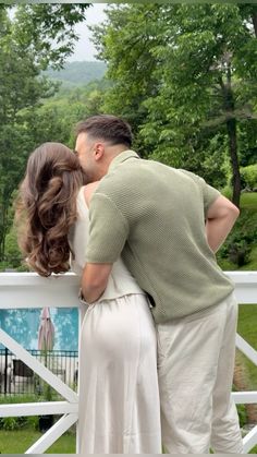 a man and woman kissing in front of a white fence with green trees behind them