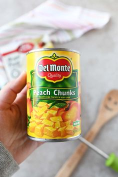 a hand holding a can of canned peach chunks next to a wooden spoon and utensils