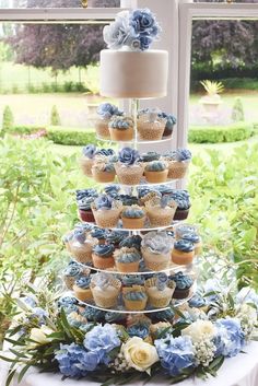 a tiered cake with cupcakes and blue flowers on the table in front of a window