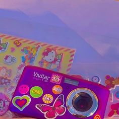 a purple digital camera sitting on top of a bed next to a box filled with stickers