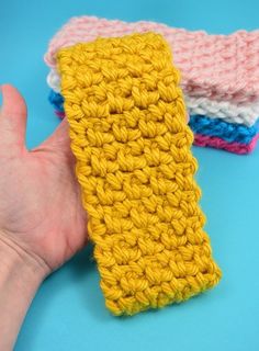 a hand is holding three crocheted items on a blue surface, one yellow and the other pink