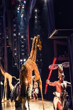 two men dressed as giraffes on stage with other performers in the background