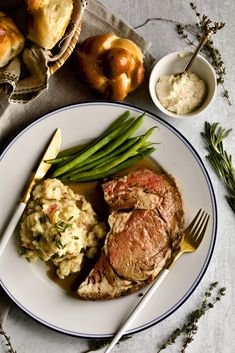 a white plate topped with meat, potatoes and green beans next to some bread rolls