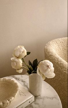 white flowers in a vase sitting on a marble table next to a book and chair