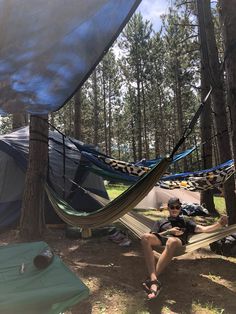 a man sitting in a hammock next to a tent on the forest floor