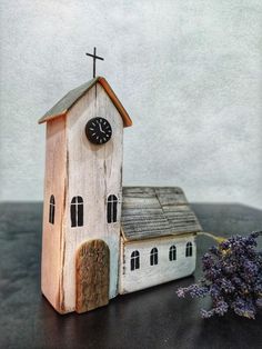 a small wooden church with a clock on the side and lavender flowers in front of it