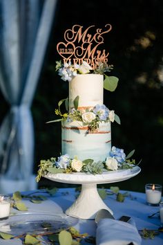 a wedding cake with blue flowers and greenery on the top is sitting on a table