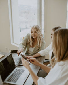 two women sitting at a table with laptops in front of them and one holding a cell phone