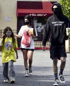 three people walking down the street with one person wearing a mask and another holding a drink