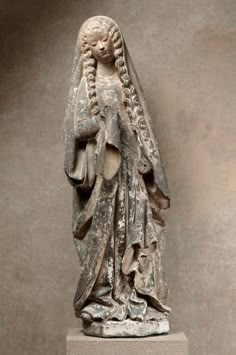 an ancient statue with long hair and flowing veil on it's head, standing in front of a gray wall