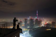 two people standing on top of a building looking at the city lights in the distance