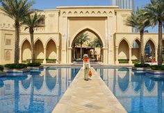 a man standing in front of a large swimming pool with palm trees around it and a tall building behind him