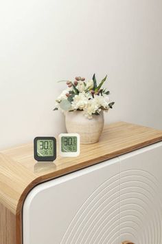 two clocks sitting on top of a wooden cabinet next to a vase with white flowers