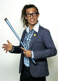 a man with long hair and glasses holds two baseball bats in one hand while smiling at the camera