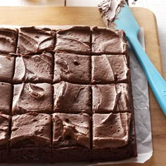 a square cake with chocolate frosting on a cutting board next to a blue spatula