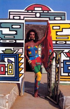 a woman standing in front of a colorful wall with an open door and red scarf hanging from it