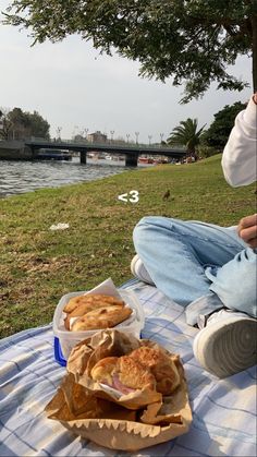 a man sitting on a blanket next to a river with some food in front of him