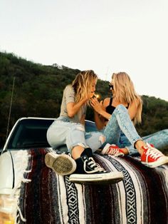 two women sitting on the hood of a car talking to each other and eating food