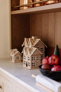 some small houses sitting on top of a counter next to apples and a bowl of fruit