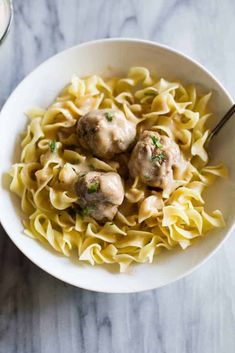 a white bowl filled with meatballs and noodles on top of a marble table next to a glass of milk