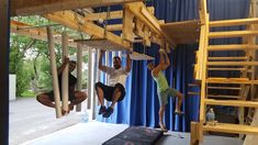 three people are hanging upside down in a room with blue curtains and wooden stairs, while one person is on a black mat