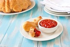 two white plates topped with pastries next to a bowl of tomato sauce and bread