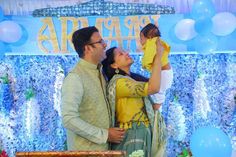 a man and woman holding a baby standing in front of a blue backdrop with balloons