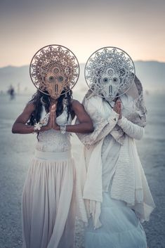 two women with masks on their faces standing in the desert at sunset, one wearing a white dress