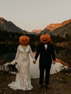 a bride and groom holding hands with carved pumpkins on their heads in front of a mountain lake