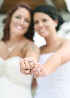 two brides holding their wedding rings and pointing at the camera with both hands together