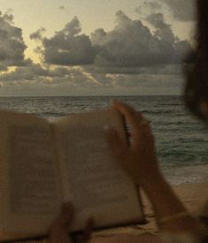 a woman is reading a book on the beach at sunset with clouds in the background