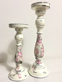 two white candlesticks with pink flowers on them sitting next to each other in front of a white wall