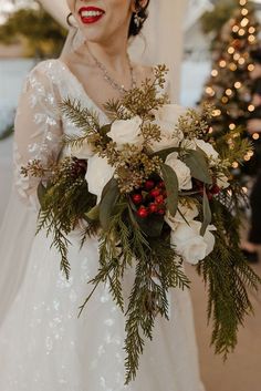 a woman in a wedding dress holding a bouquet of flowers and greenery with christmas lights behind her