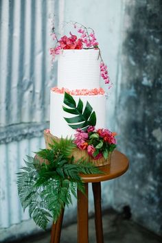 a three tiered cake with pink flowers and green leaves on top sitting on a wooden table