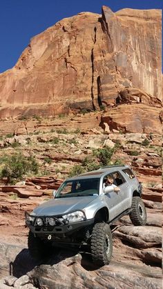 a silver truck driving on rocks in front of a large rock formation with a mountain behind it