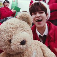 a boy in red jacket sitting next to a teddy bear with other people behind him