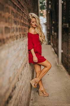 a woman leaning against a brick wall wearing sandals and a red off the shoulder dress