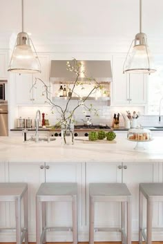 a white kitchen with four stools in front of the island and three lights hanging from the ceiling